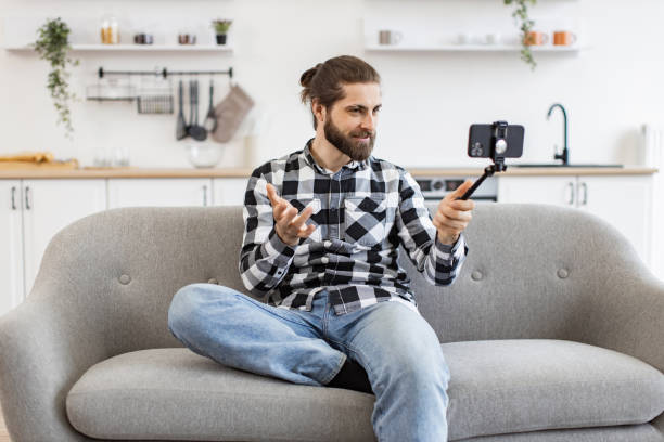 istock Young man holding phone and selfie stick while talking via webcam on kitchen. 2096210511