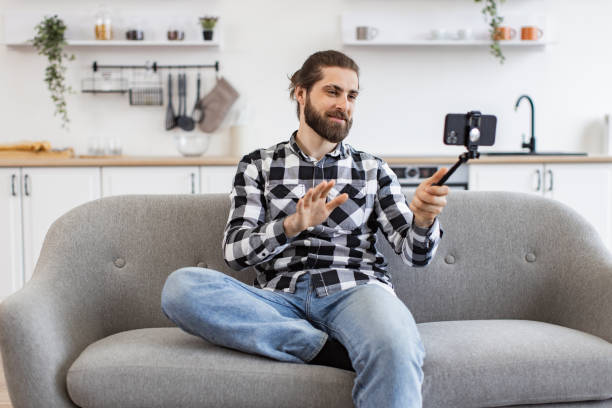 istock Young man holding phone and selfie stick while talking via webcam on kitchen. 2096210513