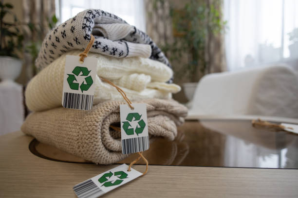 Tags with green recycling arrows on clothes folded on a table in the room. Using environmentally friendly clothing made from natural materials. Selection of old clothes for recycling.