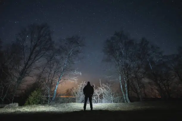 Silhouette of a man with a headlamp with a rifle behind his back in a night forest under a starry sky.High quality photo