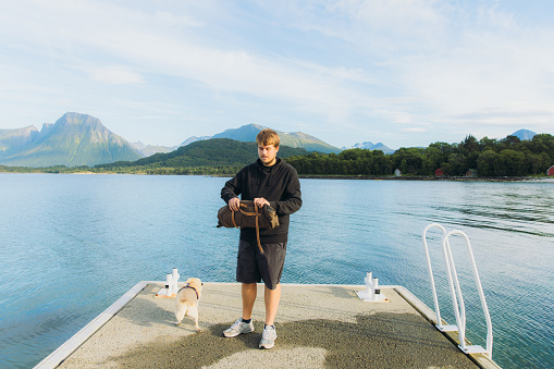 Male setting the picnic blanket on the pier with his dog enjoying summertime by the sea with idyllic mountain view in Western Norway, Scandinavia