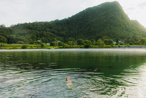 Smiling female with long hair swimming in the fjord with idyllic sunset Mountain View at the beautiful beach of in Møre og Romsdal, Norway