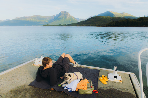 Side view of a male in black hoodie sitting on the pier with his pug enjoying summertime by the sea with mountain view in Western Norway, Scandinavia