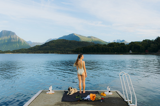 Rear view of female in swimsuit relaxing on the pier with her cute pug by the ocean during summertme with mountain view in Western Norway, Scandinavia