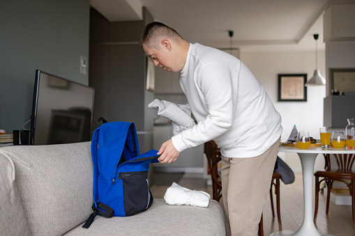 Young man with special needs packing his clothing into a backpack at home.