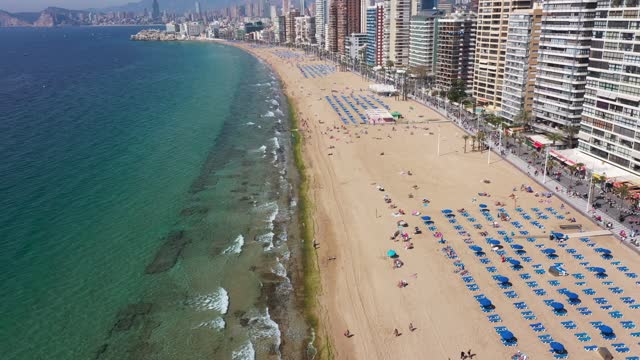 Aerial drone footage of the town of Benidorm in Spain in the summer time showing high rise apartments and building along side the Levante Beach and mountings in the background.