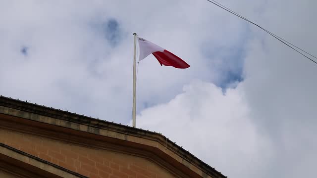 Flag of island of Malta flies against background of blue sky with white clouds.