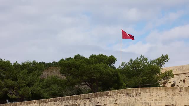 Flag of island of Malta flies against background of blue sky with white clouds.