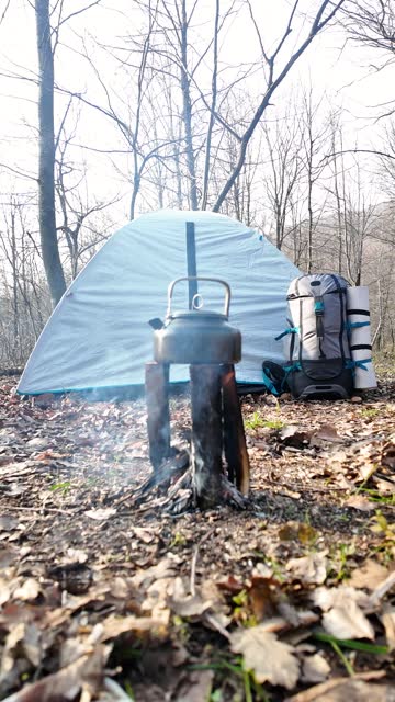Camping tent, camping backpack, camping fire and a teapot on fire in a forest. Vertical.