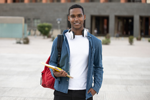 Handsome student wearing a backpack and holding a notebook looking at camera.Relaxed and happy man standing outside with headphones ready for college. High quality photo