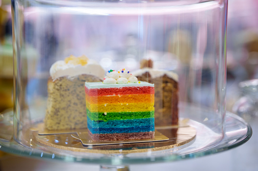 A striped rainbow cake with white frosting is displayed on a cake stand and covered by a heavy glass cover.