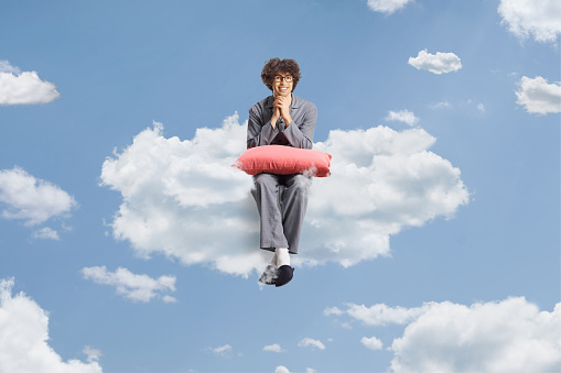 Young man in pajamas sitting on a cloud in the sky and smiling