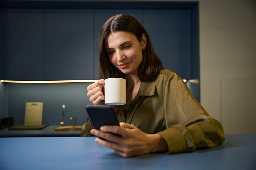 Cheerful woman writing using smartphone while having coffee break in office