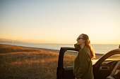 Woman enjoying sunset over the sea on the beach near her car. Travel, road trip and tourism concept.