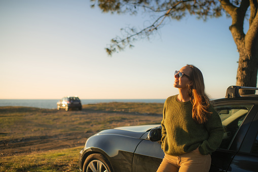 Woman enjoying sunset over the sea on the beach near her car. Travel, road trip and tourism concept.