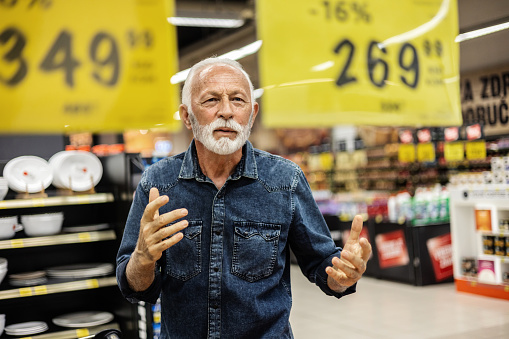 Mature man standing among the produce aisle at the supermarket and feeling upset about the increase in food prices.