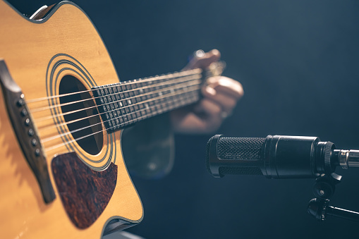 Male musician playing acoustic guitar behind microphone in recording studio. The concept of music recording, rehearsal or live performance.