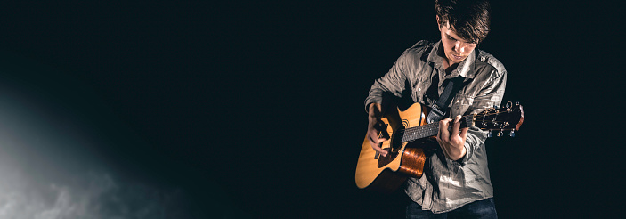 A male guitarist plays an acoustic guitar on a dark stage background. Web, social media banner template. Copy space.
