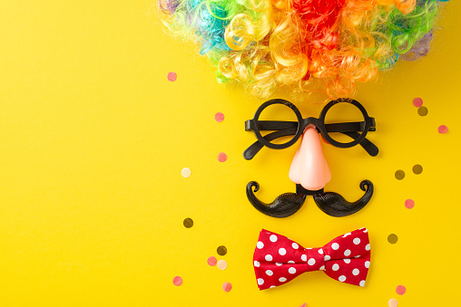 April Fool's top view setup with confetti, goofy glasses, a colorful wig, bow tie, and a fake nose, creatively placed to mimic a funny face on a vivid yellow backdrop, leaving space for your message