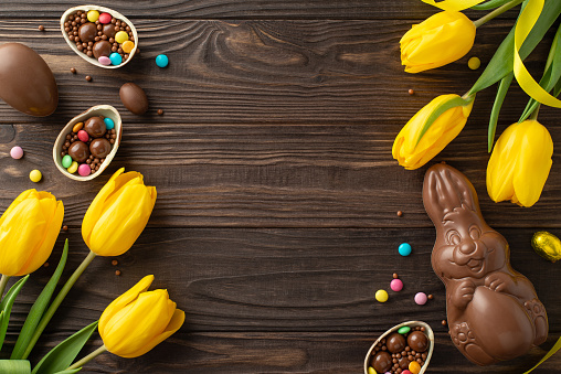 Lovely Easter snapshot: Overhead view of chocolate eggs cracked to unveil multi-hued candies, with a chocolate rabbit and tulips positioned on a wooden table, space available for text or promotions