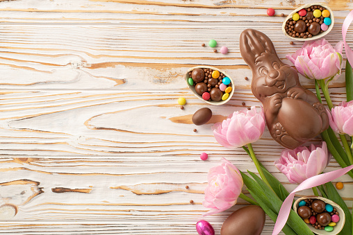 Lovely Easter setup captured from the top view, displaying shattered chocolate eggs overflowing with colorful confections, a chocolate bunny, fresh tulips on wood background, with space for messaging