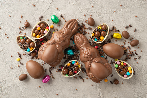 Sleek Easter sweet concept. From top view, cracked chocolate eggs full of colorful candy, a chocolate rabbits, sugar sprinkles arranged on a grey textured concrete surface