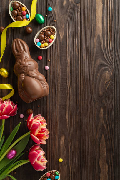 istock Festive Easter presentation: Vertical top view of shattered chocolate eggs filled with rainbow candies, alongside a sugary chocolate hare and blooming tulips on a timber backdrop, space for copy 2096635316