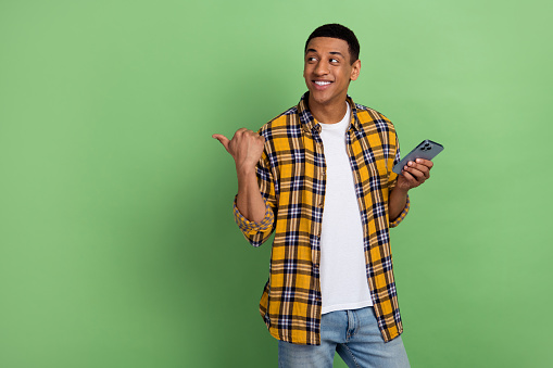 Photo of cheerful guy in yellow plaid shirt pointing finger website he created logo design using phone isolated on green color background.