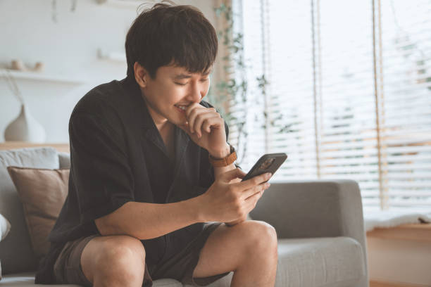 istock Happy young asian man using at smartphone in living room at home, Browsing Internet or messaging with friends, Enjoying weekend 2096683717