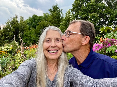 Active senior couple enjoying being together outdoors  as he kisses her on cheek while taking selfie against sky, flowers and trees