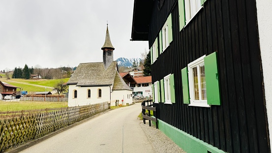 Oberstdorf, Germany - March, 10th - 2024: typical house with green shutters and a chapel in the background. Seen in the hamlet Kornau.