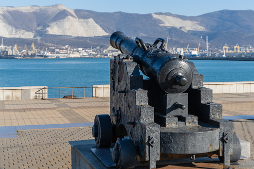 Embankment of Admiral Serebryakov. Old Ship's cannon stands on pedestal against blurred background of berths of Novorossiysk Commercial Sea Port. Novorossiysk, Russia - December 20, 2022
