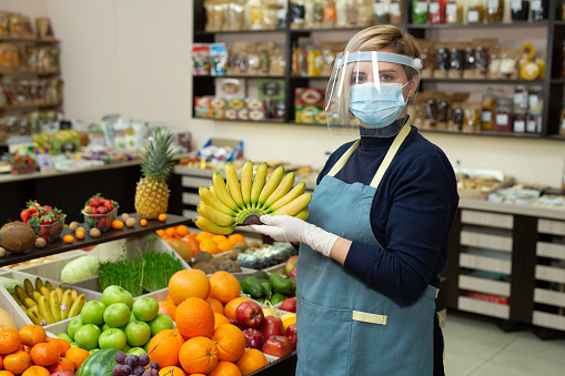 Close-up portrait of a happy, smiling female grocery store worker wearing a protective mask and gloves, holding bananas and looking at the camera.