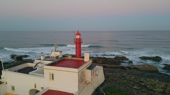 Red beacon placed on ocean shore washed by stormy waves drone view. Beautiful lighthouse standing on sea coast summer evening. Foamy swell crashing on gloomy rough rocky shoreline in super slow motion