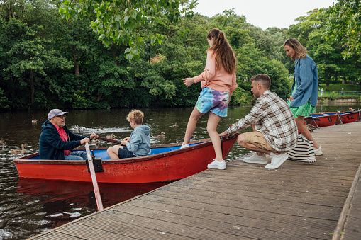 A family enjoys a day by the river, they are all wearing casual clothing in Morpeth Park in the North East of England. The Grandfather and Grandson are on a red rowing boat by the dock, the Father is on the dock steadying the boat to allow his Daughter to step onto it. The Mother is also on the dock, simply observing.