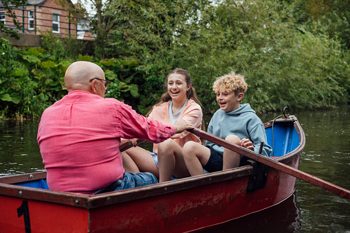 A full shot of a Grandfather with his Grandaughter and Grandson rowing along a river in Morpeth Park in the North East of England. The rowboat is red, they are all wearing casual clothing. The Grandchildren sit across from the grandfather laughing, only the back of the grandfather's head can be seen as he rows the oar.