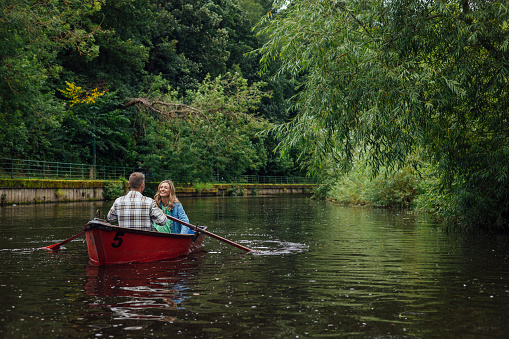 A wide shot of a Married Middle-aged couple rowing along a river in Morpeth Park in the North East of England. The couple are on a red boat, the woman smiles at her partner.