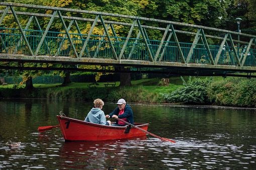 A wide-angle shot of a Grandfather rowing with his Grandson along a river in Morpeth Park in the North East of England.  The boat is red and the subjects are wearing casual clothing. The Grandfather is rowing his grandson towards the nearby bridge, a duck is floating nearby.