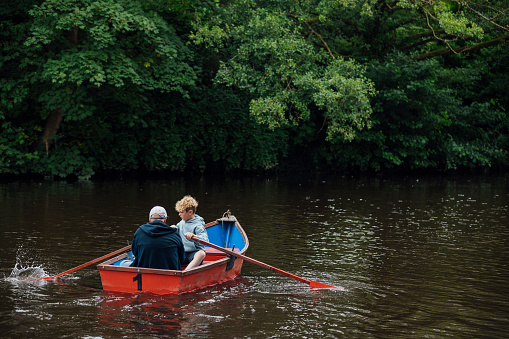 A wide-angle of a man rowing with a young boy along a river in Morpeth Park in the North East of England. The water is calm, the boy rows the red boat under the man's supervision. They both are wearing casual clothing and the man is sporting a cap but his face is out of shot.