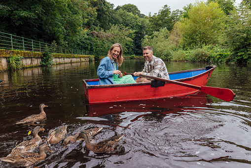 A wide shot of a Married Middle-aged straight couple rowing along a river in Morpeth Park in the North East of England. The couple are on a red boat admiring the ducks. They are both smiling and wearing casual clothing.