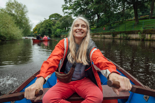 A medium torso shot of a Solo older woman sitting in a row-boat wearing red trousers and a neon orange jacket, rowing along a river in Morpeth Park in the North East of England.