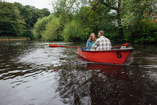 A wide shot of a Married Middle-aged couple rowing along a river in Morpeth Park in the North East of England. The couple are on a red boat, the woman smiles at her partner.