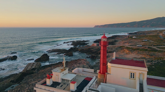 Lighthouse rising evening coast washed by beautiful foamy ocean aerial view. Red beacon standing on rocky seashore at summer twilight. Picturesque sea waves crashing of coastline at scenic sunset.