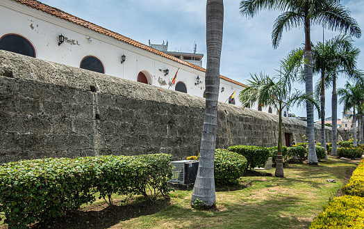 Cartagena, Colombia - July 25, 2023: Alcaldia Mayor de Cartagena de Indias, Major's office building, behind ancient rampart wall under blue cloudscape. Green trees and plants,