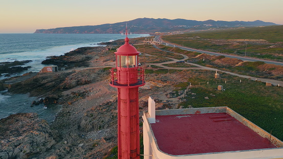 Drone view lighthouse tower placed on picturesque seashore at summer evening. High red beacon rising on rocky coast washed by beautiful atlantic ocean waves. Tranquil marine landscape at twilight.