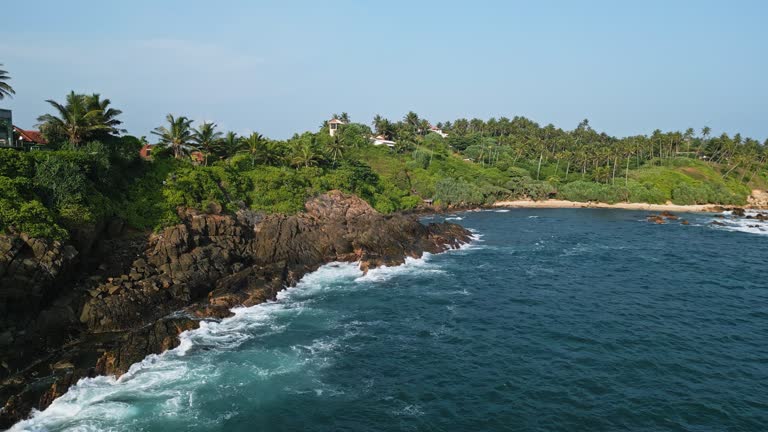 Aerial perspective lighthouse perched atop hill, adjacent to rocky shoreline with waves crashing below. Coastal beacon serves as navigation point for sea vessels, lush, tropical flora. Conveys remote tranquility, maritime safety, natural beauty, visual