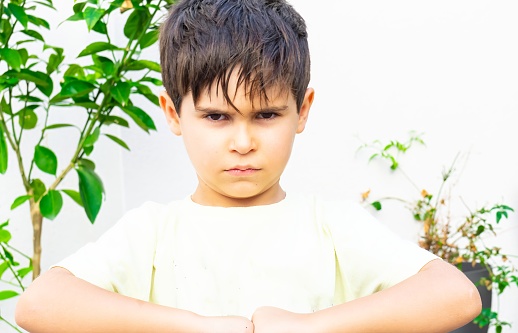 Frontal portrait of a 7-year-old Caucasian boy with angry expression in the garden. Concept of child behavior. Horizontal.