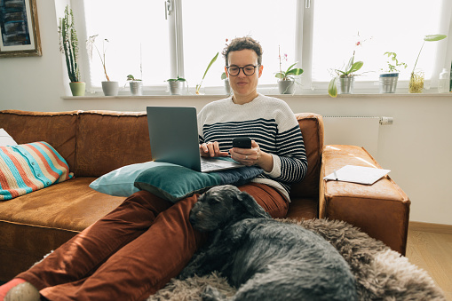 A woman is sitting on a couch with a laptop in front of him. A dog is laying on a rug next to him