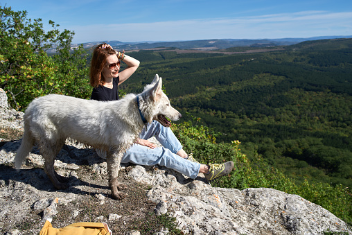 Free happy woman sitting resting and enjoying nature with white dog in a top of mountains above valley forest. Healthy lifestyle concept.