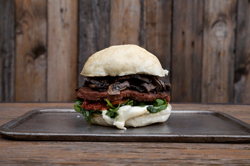Vegan cuisine. Plant based food. Closeup view of a Not burger with potato bread, mushrooms, sun dried tomatoes, arugula and NotCo mayonnaise, with a rustic wooden background